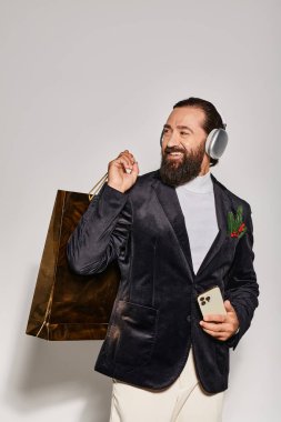 Kablosuz kulaklıklı neşeli sakallı adam gri arka planda akıllı telefon ve alışveriş çantası tutuyor.