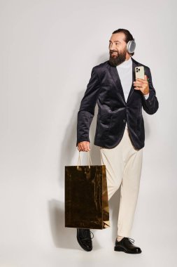 Kablosuz kulaklıklı, gülümseyen sakallı adam gri arka planda akıllı telefon ve alışveriş çantası tutuyor.