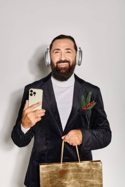 Kablosuz kulaklıklı neşeli sakallı adam elinde akıllı telefon ve gri arka planda hediye çantası tutuyor.