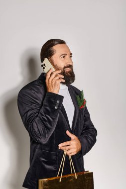 Takım elbiseli mutlu sakallı adam akıllı telefonla konuşuyor ve elinde gri arka planda Noel hediyesi çantası tutuyor.