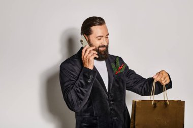 happy bearded man in suit talking on smartphone and holding Christmas gift bag on grey backdrop clipart