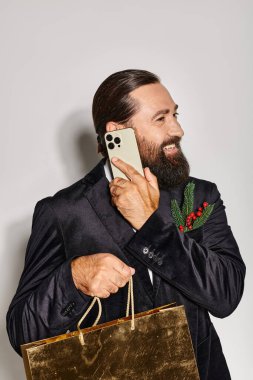 Takım elbiseli mutlu sakallı adam akıllı telefonla konuşuyor ve elinde gri arka planda Noel hediyesi çantası tutuyor.