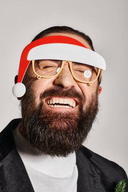 joyous bearded man in festive glasses with santa hat posing in suit and smiling on grey backdrop clipart