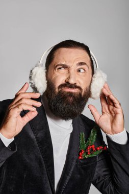 emotional bearded man in suit with Christmas spruce in pocket wearing ear muffs on grey backdrop clipart