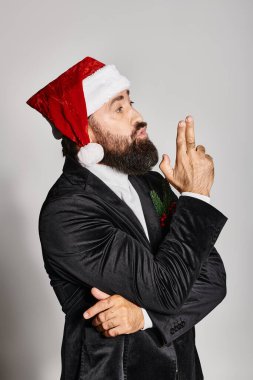 Resmi giyinmiş sakallı yakışıklı bir adamın portresi ve Noel Baba şapkası gri arka planda poz veriyor.