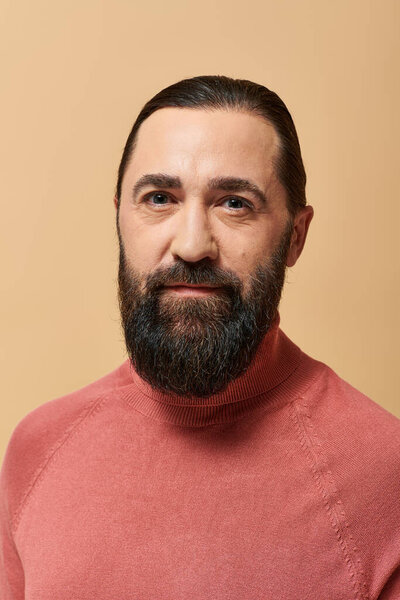 portrait, serious and handsome man with beard posing in pink turtleneck jumper on beige background