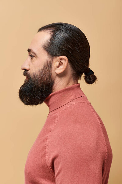 side view, handsome serious man with beard posing in pink turtleneck jumper on beige backdrop