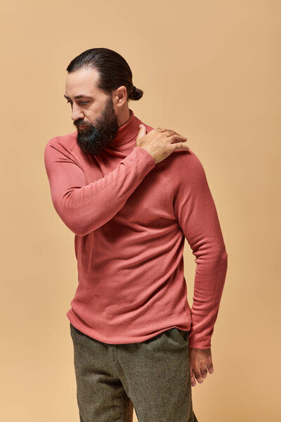 portrait, serious and handsome man with beard posing in pink turtleneck jumper  on beige background