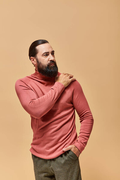 portrait of serious handsome man with beard posing in pink turtleneck jumper on beige background