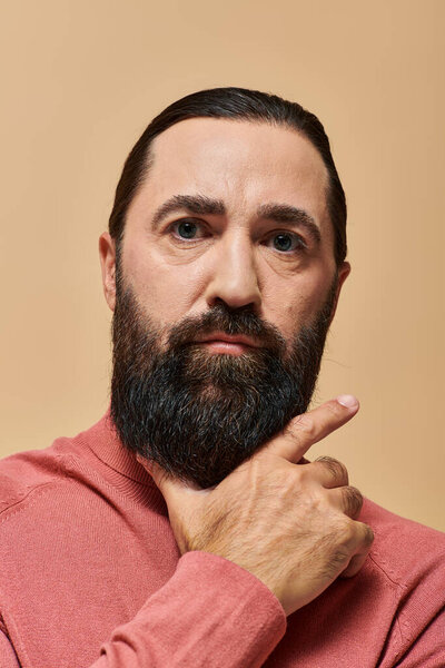 portrait of serious good looking man with beard posing in pink turtleneck jumper on beige backdrop