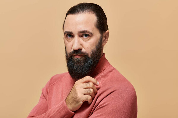 portrait of good looking serious man with beard posing in pink turtleneck jumper on beige backdrop
