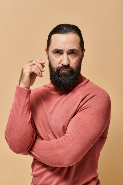 serious and handsome man with beard posing in pink turtleneck jumper on beige background, portrait