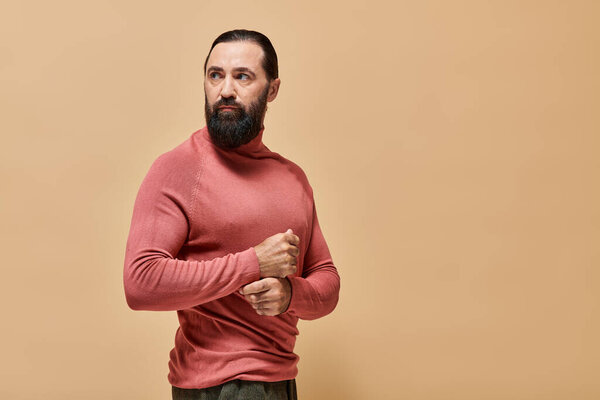 good looking and serious man with beard posing in pink turtleneck jumper on beige backdrop, portrait