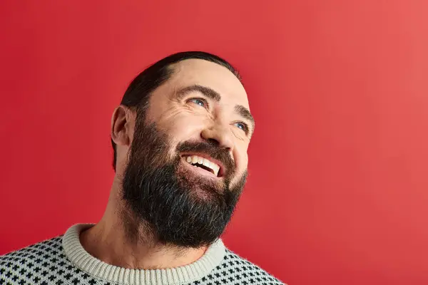 stock image happy bearded man in winter sweater with ornament smiling on red backdrop, Merry Christmas