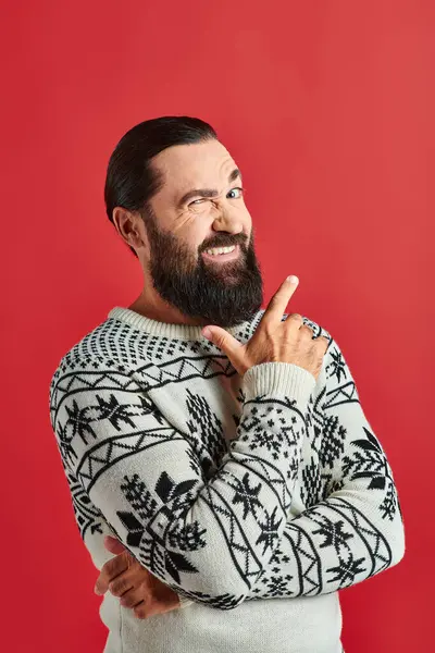 Stock image cheerful bearded man in winter sweater with ornament pointing on red backdrop, Merry Christmas