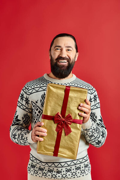 bearded joyful man in winter sweater with ornament holding Christmas present on red background