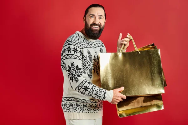 stock image happy bearded man in sweater with ornament holding shopping bags on red backdrop, Christmas present