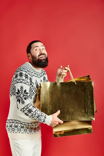 stock image happy bearded man in sweater with ornament holding shopping bags on red backdrop, Christmas present