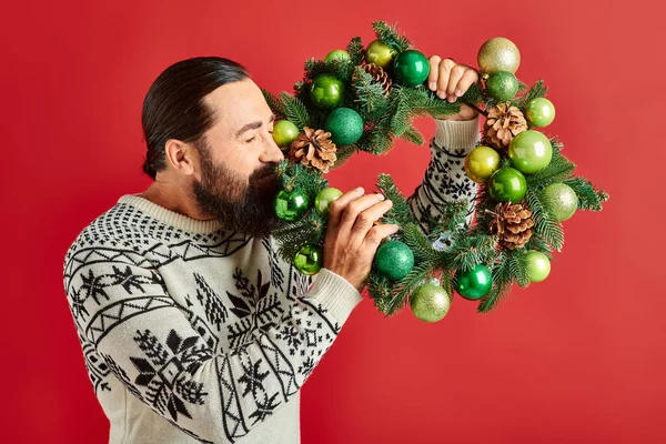 冬のセーターで幸せなひげを被った男 赤い背景 クリスマスに問題を抱えた装飾的な羽根を保持 — ストック写真