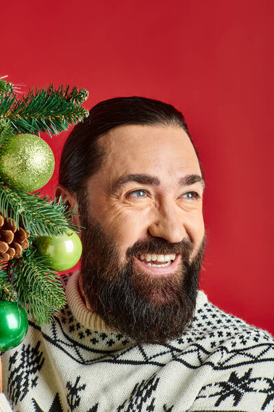 joyful bearded man in Christmas sweater holding decorated wreath with baubles on red backdrop
