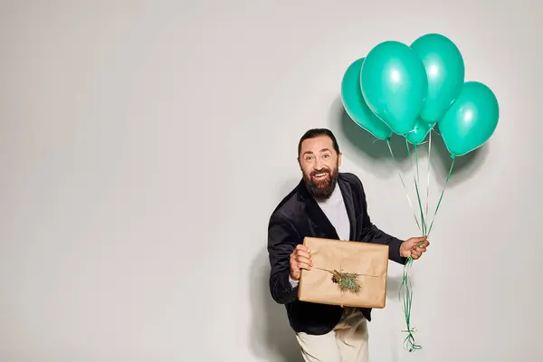 stock image happy bearded man in suit holding blue balloons and wrapped Christmas present on grey backdrop