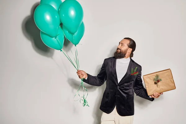 Stock image happy bearded man in formal attire holding blue balloons and Christmas  present on grey backdrop