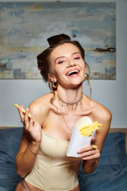 cheerful young woman in sexy beige corset with hair curlers posing with fries and smiling at camera clipart
