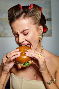 Saç bigudileri ve aksesuarları olan genç ve güzel bir kadın iştahlı lezzetli bir burger yiyor.