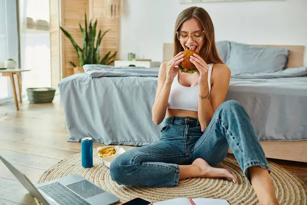 cheerful attractive woman in casual attire sitting on floor while working and enjoying burger