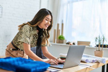 Gülümseyen Asyalı moda tasarımcısı atölyede çalışıyor. Laptop ve kumaş örnekleri, küçük işletmeler.