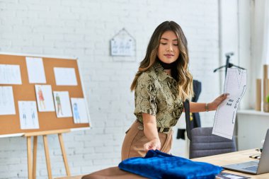 Yetenekli Asyalı moda tasarımcısı dikiş deseni tutuyor ve kendi atölyesinde kumaş örneğine dokunuyor.