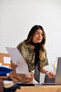 talented asian designer with sewing patterns looking at laptop in private workshop, small business clipart