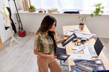 young asian designer looking at color swatches near laptop with blank screen in fashion atelier clipart