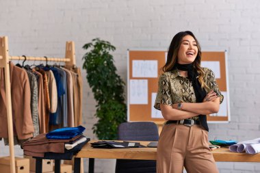 heyecanlı stil Asyalı tasarımcı kollarını kavuşturmuş özel moda atölyesinde çalışma masasının yanında gülüyor.