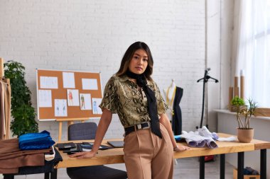 Moda stüdyosundaki çalışma masasının yanındaki kameraya bakan çekici Asyalı stilist.
