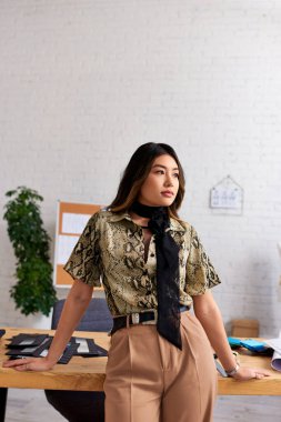 Düşünceli Asyalı kıyafet tasarımcısı kişisel moda stüdyosunda çalışma masasının yanından uzağa bakıyor, yaratıcılık