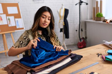 Genç Asyalı kıyafet tasarımcısı özel atölyedeki çalışma masasında kumaş örneklerinin kalitesini kontrol ediyor