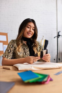 happy asian dressmaker holding quality threads near fabric and sewing supplies in personal atelier clipart