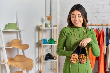 smiling asian clothes designer showing stylish animal print shoes in private atelier, small business clipart