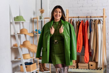 Moda stüdyosunda moda kıyafetleri ve yeşil deri ceketle poz veren neşeli Asyalı stilist.