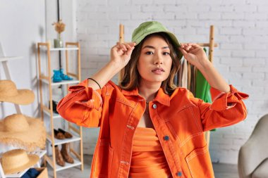 Parlak turuncu elbiseli, panama şapkalı ve atölyedeki kameraya bakan Asyalı bir stilist.