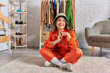Panama şapkalı ve turuncu elbiseli yaratıcı Asyalı tasarımcı özel moda stüdyosunda yerde oturuyor.