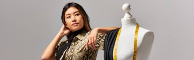 modern asian stylist looking away near mannequin with fabric and measuring tape on grey, banner clipart