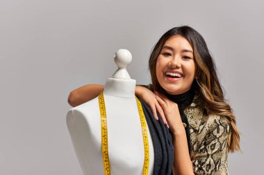 excited asian stylist smiling at camera near mannequin with fabric and measuring tape on grey clipart