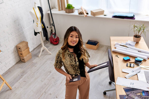 весёлый азиатский дизайнер с цветными образцами, смотрящий на камеру в современном ателье, с высоким углом обзора