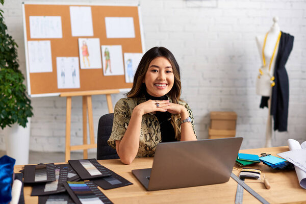 cheerful asian stylist looking at camera near laptop and color swatches on work desk in atelier