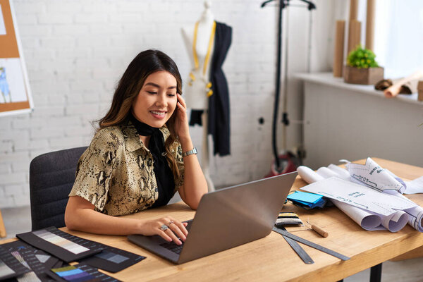 happy asian fashion designer working on laptop near sewing pattens and swatches in own atelier