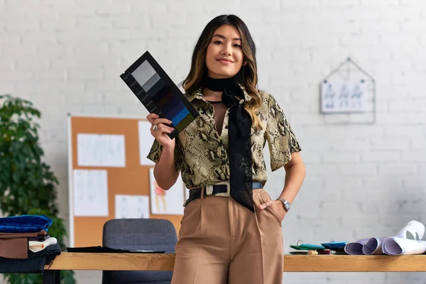 stock image joyful confident asian designer with color samples and hand in pocket looking at camera in atelier