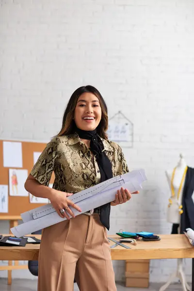 stock image joyful successful asian designer with sewing patterns looking at camera in private atelier