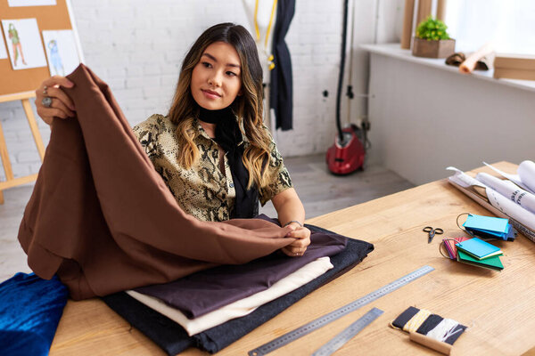 young creative asian stylist looking at fabrics samples in own atelier, modern fashion business